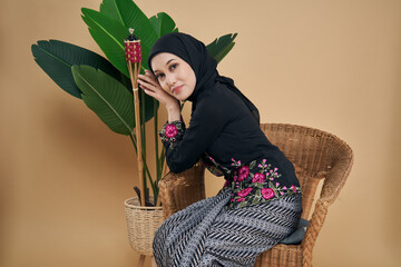Beautiful Asian model wearing black kebaya dress with hijab, sitting on a rattan chair with bamboo...