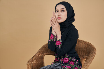 Beautiful Asian model wearing black kebaya dress with hijab, sitting happily on a rattan chair isolated over beige background. Eidul fitri festival, fashion and beauty concept.