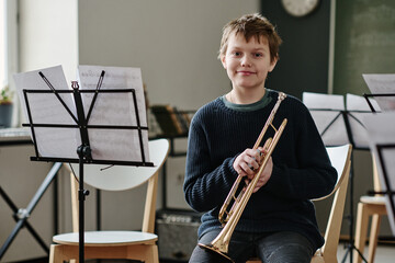 Medium portrait of cheerful Caucasian teen boy holding trumpet sitting in classroom equipped with...