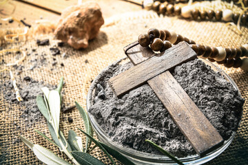 Detail of glass container with ashes and wooden cross on burlap cloth with olive leaves on a wooden table. Elevated view.