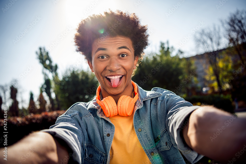 Canvas Prints Photo of funky carefree childish youngster dressed jeans jacket with headphones on neck making selfie stick out tongue in park outdoors