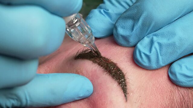 Microblading tattoo , permanent make-up. The master in gloves, using a special needle, injects pigment into the skin and colours the eyebrows with the hair technique, close-up view