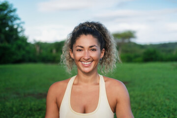 Portrait of a young attractive Latino athletic woman who does yoga against the backdrop of a green forest. Close-up of a yoga trainer, she looks at the camera and smiles. Wellness practice in nature.