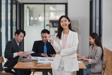 Beautiful smart Asian businesswoman standing in the office room with her teammates in the background.