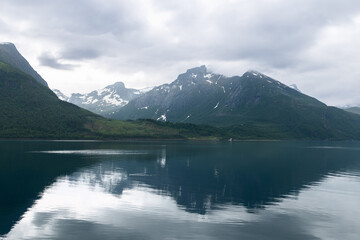 Reflective fjord waters capture the moody essence of a cloudy Norwegian day, with mountains veiled in mist creating a captivating, somber scene