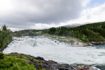 Laksforsen waterfall's raw power on display, cascading forcefully in a natural setting framed by verdant woods and mountain peaks. Norway