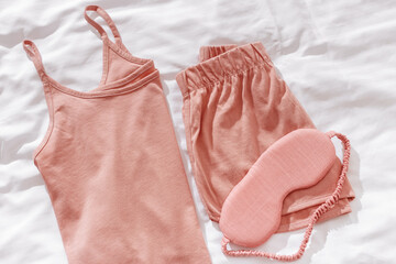 Top view pink pajama and eye sleep mask on white crumpled bedclothes. Cozy pyjamas for comfort rest at night. Flat lay from singlet, shorts, sleeping mask pastel pink color, sleep well