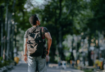 Man traveling with backpack and camera looking at park area in forest at sunny day. Solo traveller concept.