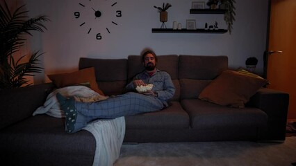 Front view of a young Caucasian man having fun and laughing watching TV while lying on the sofa at home at night eating popcorn. Male enjoying a movie in the living room in the dark.