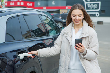 Young woman charging her electric car at the gas station, using a smartphone. Eco fuel concept. The concept of environmentally friendly transport. Recharging battery from charging station.