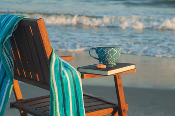 Antique beach chair with book towel and coffee cup sits on the beach at sunrise