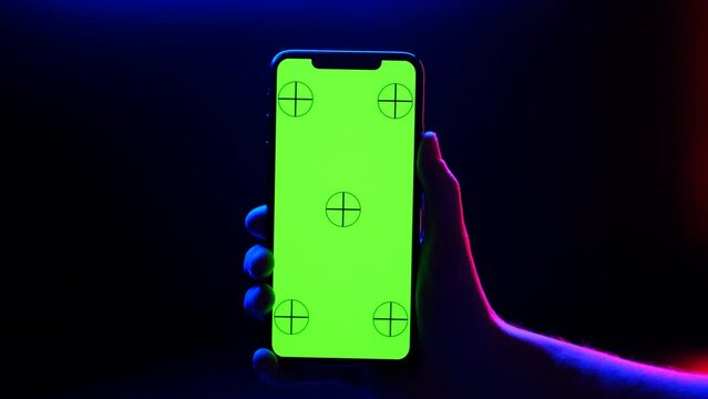 Mobile phone in hand. Holding smartphone with red blue neon lighting on dark background. Green chromakey screen with markers. Hand lifts phone up and holds it still.