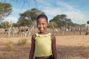 african village child on a dirt road , kraal with small livestock grazing in the background,