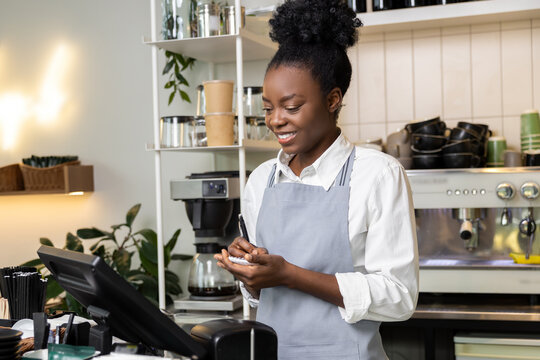 Woman bartender in coffee shop wearing apron writing order in notebook serving for client