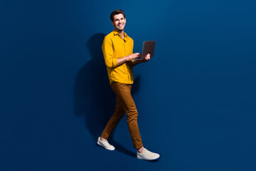 Full body photo of attractive young guy walking hold netbook smm manager wear trendy yellow clothes isolated on dark blue color background