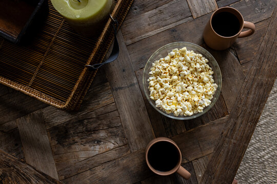A bowl of popcorn sits on a rustic wooden table next to two brown mugs