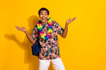 Photo of young funky positive guy in exotica style flowers necklace and backpack tourist traveler isolated on yellow color background