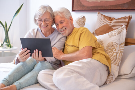 Video call concept. Portrait of happy senior couple communicating in remote chat with family and friends - Grandparents with digital tablet sitting in bed, wireless technology