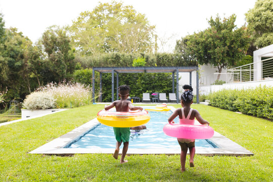African American brother and sister holding colorful floaties approach a pool with copy space