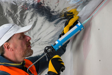 a worker in a vest, cap and gloves uses a vapor barrier glue gun
