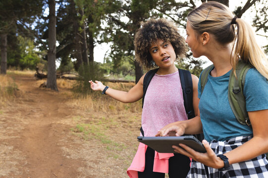 Young biracial woman with curly hair gestures while holding a tablet, discussing with a young Caucas