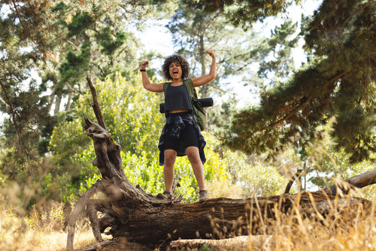 Young biracial woman stands triumphantly on a fallen tree trunk outdoors on a hike