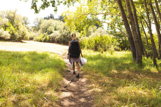 A young woman walks along a dirt path in a sunny, wooded area, collecting trash