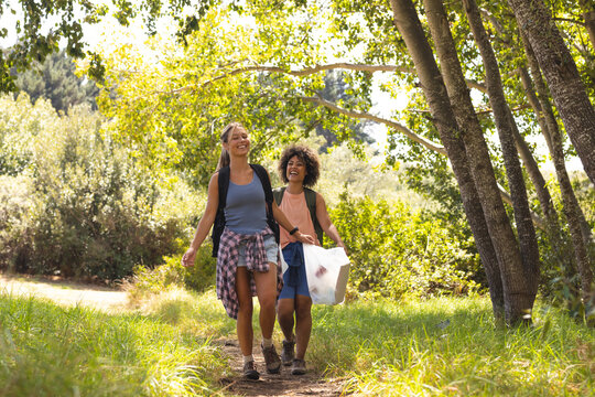 Two women are walking on a forest trail, one with blonde hair, the other with curly dark hair, colle