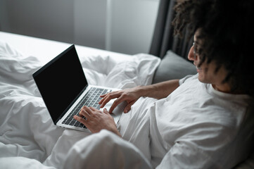 Brunette man in white tshirt lying in bed and working on laptop