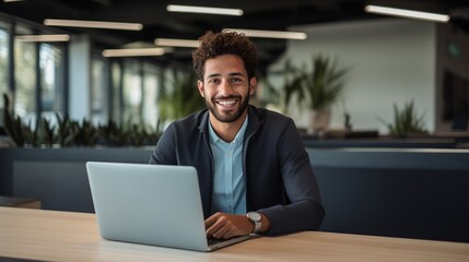 Capture the essence of a good-looking millennial office employee or student sitting at a desk in front of a laptop, smiling, and confidently looking at the camera.  - obrazy, fototapety, plakaty