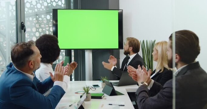 Mixed race businesspeople man and woman looking on big green screen chroma are satisfied of results of joint work and clapping hands simultaneously. Business technology concept.