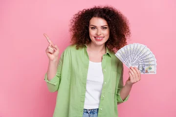  Photo portrait of lovely young lady hold banknotes fan point empty space dressed stylish green garment isolated on pink color background © deagreez