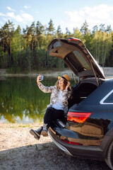 Selfie in nature. Happy woman sits in the trunk of a car and uses a smartphone. The concept of traveling by car, active lifestyle, blogging.