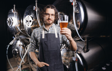 Expert brewer in apron holds glass of craft beer and checking quality and color. Worker man sommeliers taste drink on brewery factory