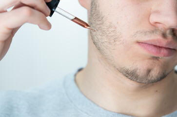young man applying beard growth oil with pipette close up. Close-up image of handsome man holding...