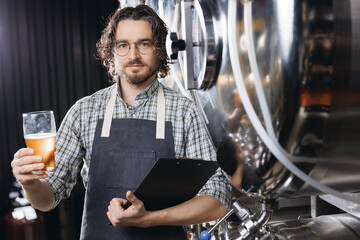 Owner brewery, bearded hipster man in apron hold glass and control quality of craft beer from tank