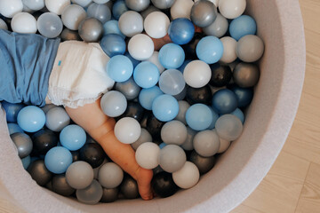 Happy little boy play in dry pool with colorful balls in living room, aerial top view. Concept lifestyle childhood moment