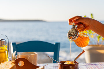 Close up of hand with honeypot pouring honey over yogurt on a table with full breakfast in a...