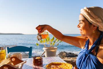 Beautiful woman in a Greek outfit enjoying a splendid breakfast while pouring honey over yogurt at...