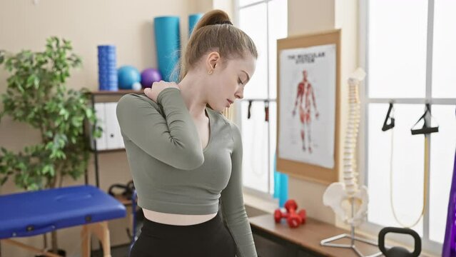 A young woman in a clinic holds her neck indicating pain, surrounded by rehabilitation equipment.