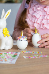 Obraz na płótnie Canvas A cute girl with pink bunny ears makes an Easter craft - decorates an egg in the form of a unicorn with rhinestones, horn, flowers in the interior of a house with plants.