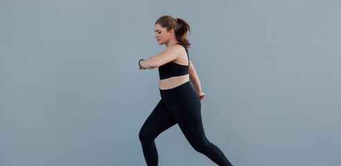 Plus-size female jogging at a grey wall. Woman with a plus-size body running outdoors and looking at her smartwatch.