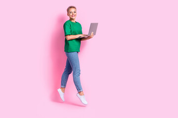 Full length photo of cheerful nice intelligent woman wear oversize t-shirt jumping with laptop in hands isolated on pink color background