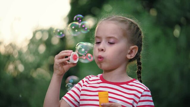 Cheerful girl with thin braids engages in whimsical activity of blowing soap bubbles. Happy cute girl with slender plaits delights in blowing soap bubbles in garden. Cute kid plays with soap bubbles