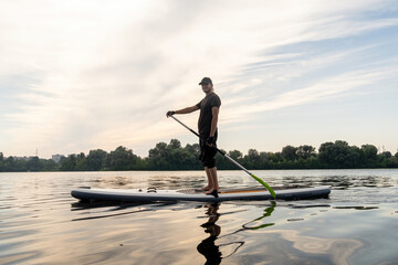 Young man leisurely glides on paddleboard across urban lake. Male devotes holiday abroad to active recreation. Improving physical health and body shape