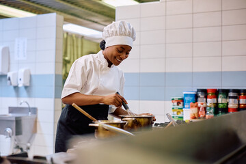 Happy black female chef cooking in kitchen in restaurant. - obrazy, fototapety, plakaty