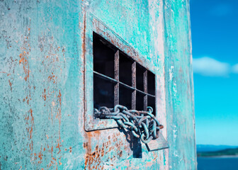 A worn and rusty grate lock and chains affixed to the window of an old house, angled shot with the sea blurred in the background. Symbolic juxtaposition of freedom and captivity.
