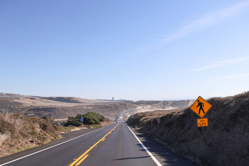 Long desert road beside mountains in the distance with crosswalk sign that reminds drivers to stay alert ensuring safety for pedestrians. Roadside call box provides a direct line to emergency services