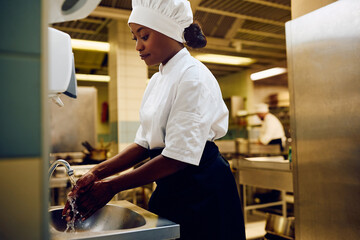 Black female chef washing hands under kitchen sink in restaurant.