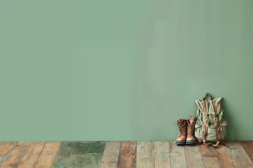 Poster Sac à dos et chaussures de randonnée à côté contre un mur vert posés sur un parquet coloré. Fond vert avec espace négatif pour texte, copyspace. Vacances vertes, nature, réservation, vacances, camping © Noble Nature